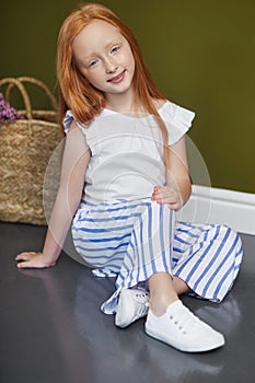 Little red-haired girl with a basket of flowers posing on an olive background. Spring portrait of a redhead girl with blue eyes.