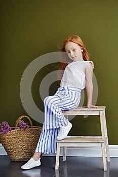 Little red-haired girl with a basket of flowers posing on an olive background. Spring portrait of a redhead girl with blue eyes.