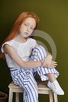 Little red-haired girl with a basket of flowers posing on an olive background. Spring portrait of a redhead girl with blue eyes.