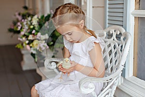 The little red-haired girl with pigtails holding a yellow chicken on a background of white houses and flowers