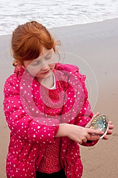 little red haired girl in winter clothes at beach holding a paua shell