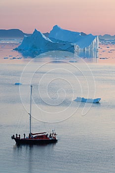 Little red sailboat cruising among floating icebergs in Disko Bay glacier during midnight sun season of polar summer. Greenland