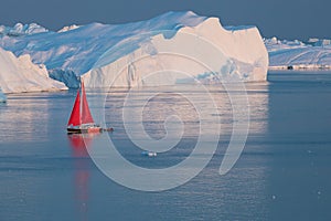 Little red sailboat cruising among floating icebergs in Disko Bay glacier during midnight sun season of polar summer. Greenland