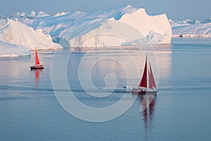 Little red sailboat cruising among floating icebergs in Disko Bay glacier during midnight sun season of polar summer. Greenland