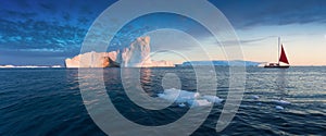 Little red sailboat cruising among floating icebergs in Disko Bay glacier during midnight sun season of polar summer. Greenland