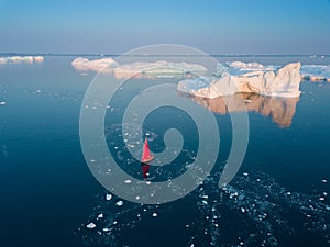Little red sailboat cruising among floating icebergs in Disko Bay glacier during midnight sun season of polar summer. Ilulissat