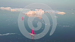 Little red sailboat cruising among floating icebergs in Disko Bay glacier during midnight sun season of polar summer. Ilulissat,