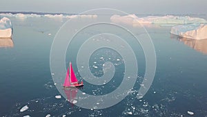 Little red sailboat cruising among floating icebergs in Disko Bay glacier during midnight sun season of polar summer. Ilulissat,