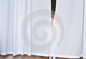 The little redhead child looks out because of white curtains