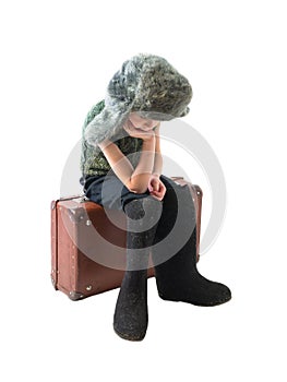 The little Russian boy in a fur hat and boots sitting on old suitcase and thinking