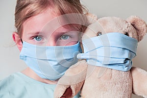 Little sad blonde girl wearing protect mask and hugging bear toy in mask. Coronavirus protecting Covid19 concept.