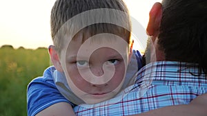 Little sad boy looking into camera and hugging his dad against background of sunset. Young father holding in hands his