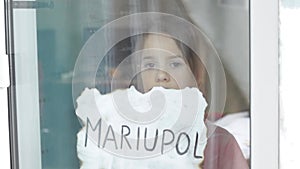 a little sad girl looks through the window holding a piece of paper with the inscription Mariupol