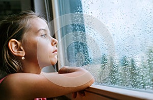 Little sad girl pensive looking through the window glass with a lot of raindrops. Sadness childhood concept image