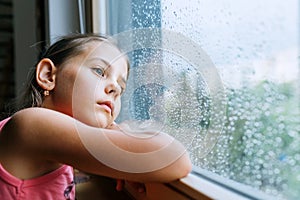 Little sad girl pensive looking through the window glass with a lot of raindrops. Sadness childhood concept image