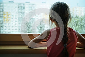 Little sad girl pensive looking through the window glass with a lot of raindrops. Sadness and loneliness childhood concept image.