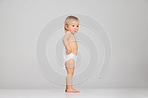 Little sad scared toddler girl, baby in diaper standing isolated on grey studio background. Concept of emotions