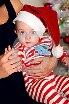 Little Santa baby posing beside Christmas tree at cozy home with New Year decoration,sitting at mom's hands