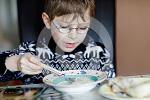 Little school boy with glasses eating vegetable soup indoor. Blond child in domestic kitchen or in school canteen. Cute