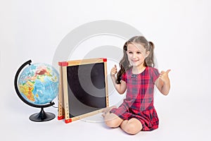 A little school girl of 7 years old sits in a red dress at a chalkboard with a globe on a white isolated background and shows the