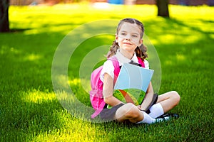 Little school girl with pink backpack sitting on grass after lessons looking side away thoughtfully , read book or study