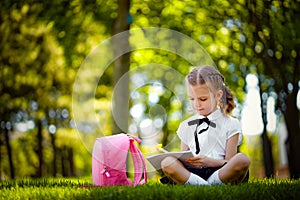 Little school girl with pink backpack sitting on grass after lessons and read book or study lessons, thinking ideas