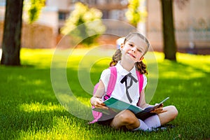 Little school girl with pink backpack sitting on grass after lessons and read book or study lessons, thinking ideas