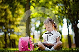Little school girl with pink backpack sitting on grass after lessons and read book or study lessons, thinking ideas
