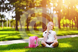 Little school girl with pink backpack sitting on grass after lessons and thinking ideas, read book and study lessons