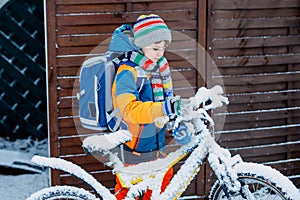 Little school kid boy of elementary class walking to school during snowfall. child removing snow from bicycle.