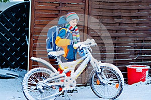 Little school kid boy of elementary class walking to school during snowfall. child removing snow from bicycle.