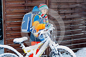 Little school kid boy of elementary class walking to school during snowfall. child removing snow from bicycle. Student