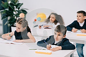 Little schoolchildren sitting at desks and writing in exercise books