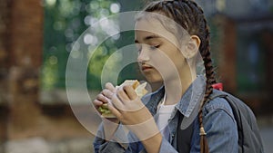 A little schoolgirl is eating a sandwich standing during the break on the schoolyard. The child stands and eats bread