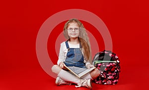 a little schoolgirl girl with a backpack is sitting cross-legged on a red  background reading a book