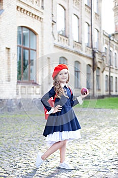 Little schoolgirl in a red beret and dress with lunch near the school. preschool child with an apple and a backpack on his first d