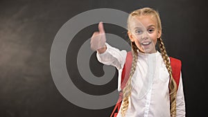 Little schoolgirl showing thumbs-up on camera against blackboard, education