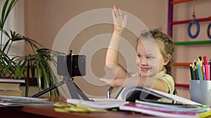 A little schoolgirl sitting at a table with a smartphone raises her hand to answer.