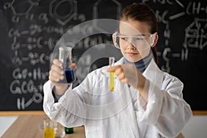 little scientist girl with glasses in lab coat with chemical flasks, on school blackboard background with science