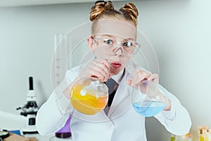 Little scientist in lab coat and protective eyeglasses making experiment with reagents in flasks