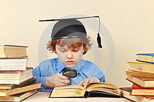 Little serious boy in academic hat studies an old books with magnifying glass