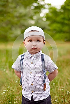 Little serious boy in suspenders and a cap hides his hands behind back in park