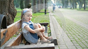 Little serious child girl sitting alone on a bench in summer park