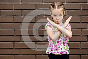 A little serious girl stands near a brick wall and shows enough or a stop sign with a gesture