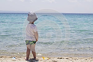 A little shy boy with hat standing in the sand near water and lo