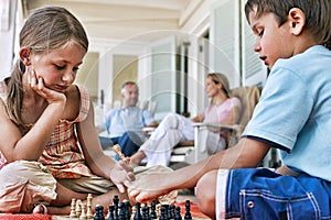 Little siblings playing chess at porch with parents sitting in background