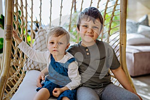 Little siblings playing, having fun and swinging on hanging chair in conservatory at home.