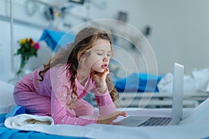 Little sick girl lying at hospital bed and using laptop.