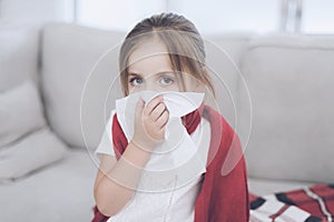 Little sick girl sits on a white couch wrapped in a red scarf. She blows her nose into a napkin