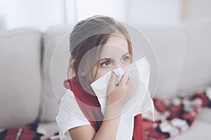 Little sick girl sits on a white couch wrapped in a red scarf. She blows her nose into a napkin